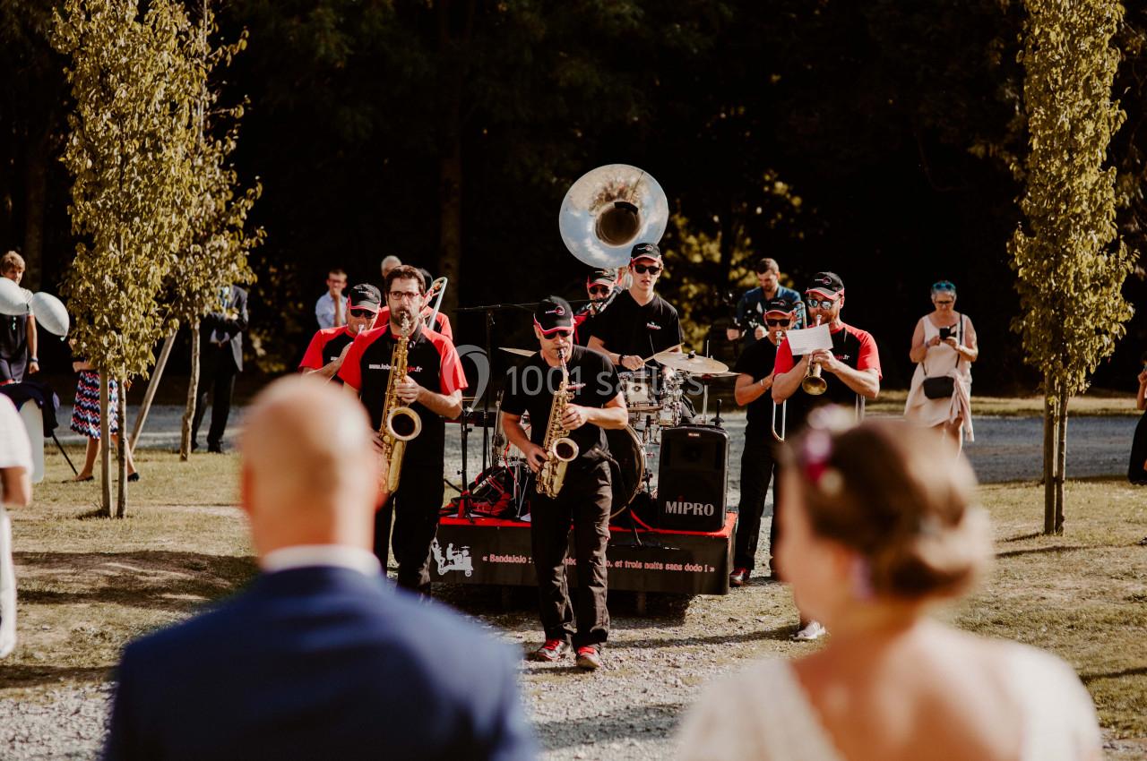 Un groupe de musiciens joue en plein air lors d'un événement, entouré d'arbres et de spectateurs.