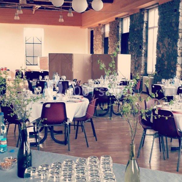Salle de réception avec des tables rondes dressées, décorée de branches dans des vases, éclairée par de grandes fenêtres.
