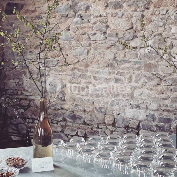 Des verres alignés sur une table avec une bouteille en guise de vase contenant des branches, devant un mur en pierre.