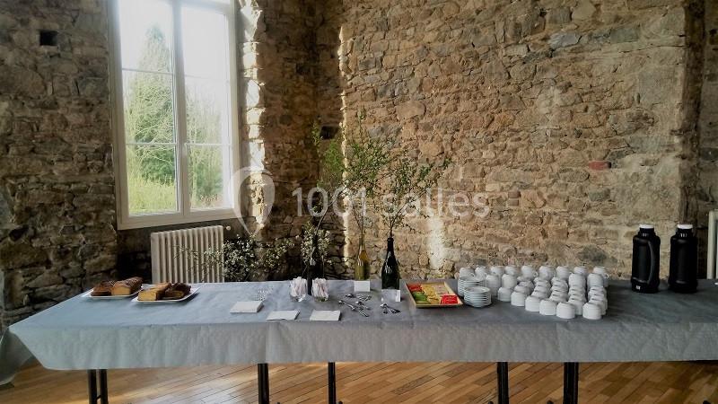 Table dressée dans une salle en pierre, avec tasses, bouteilles thermos, viennoiseries et décorations florales.