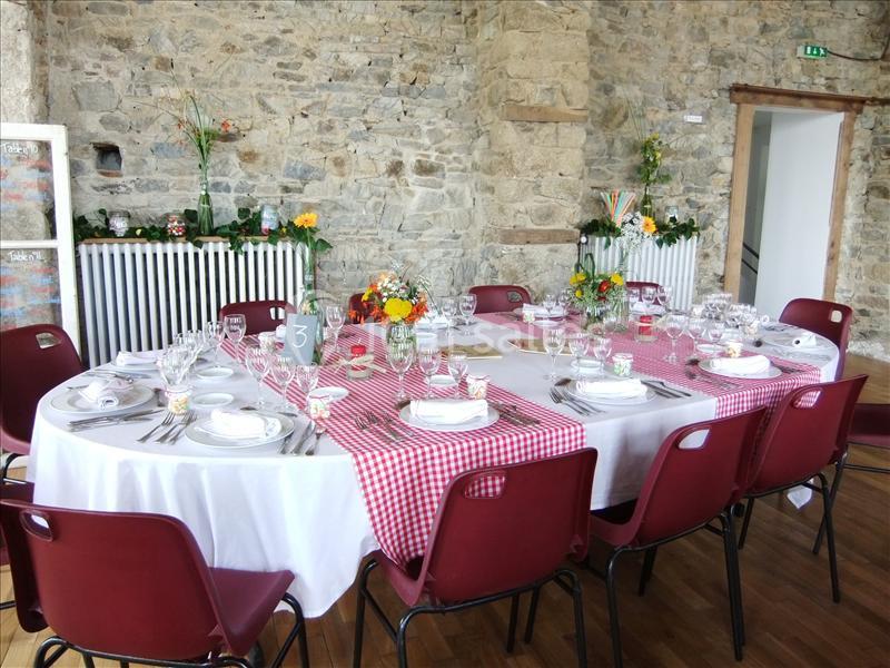 Table ronde dressée pour un repas, nappes blanches et rouges, vaisselle élégante, décor floral dans une salle en pierre.
