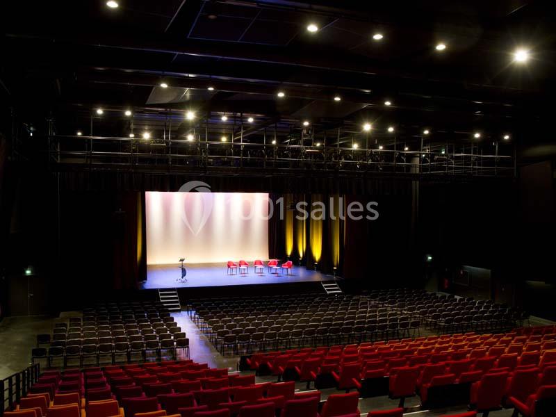 Salle de spectacle vide avec sièges rouges, scène éclairée et quelques chaises alignées sous des projecteurs.