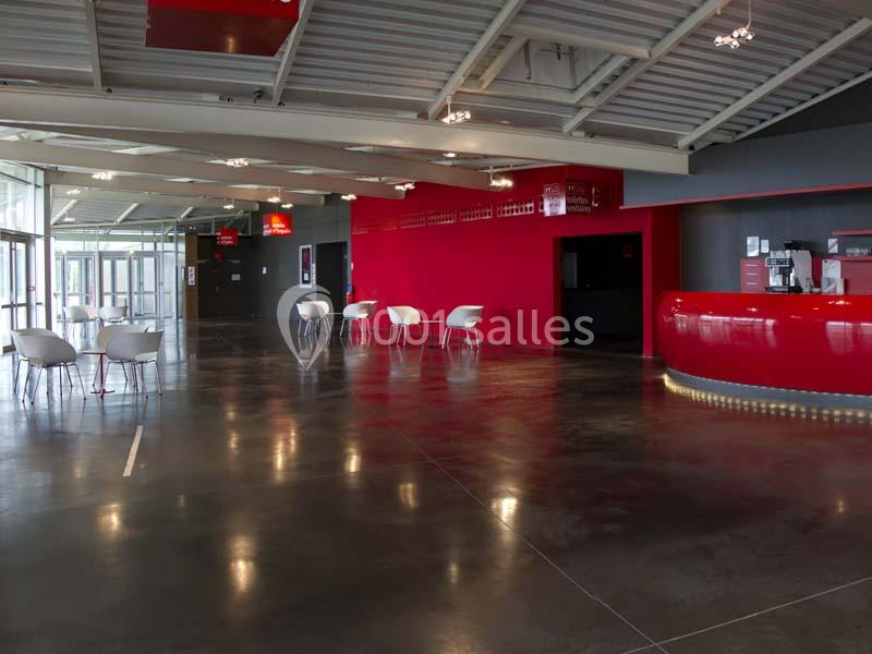 Hall moderne avec sol en béton poli, comptoir rouge, chaises blanches et lumière naturelle entrant par de grandes fenêtres.