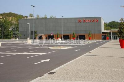 Parking vide devant un bâtiment moderne gris avec l'enseigne ’Le Dastala’ et des arbres en arrière-plan.