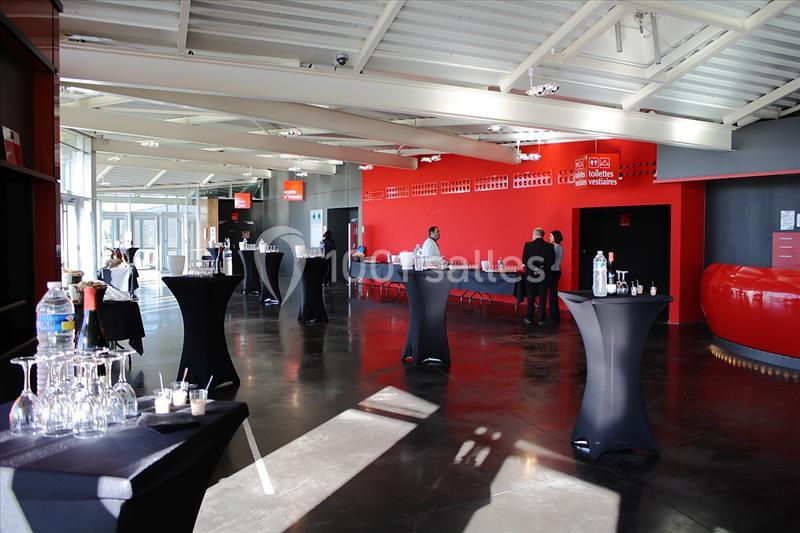 Salle moderne avec tables hautes noires, verres disposés, et quelques personnes dans un espace lumineux aux murs rouges.