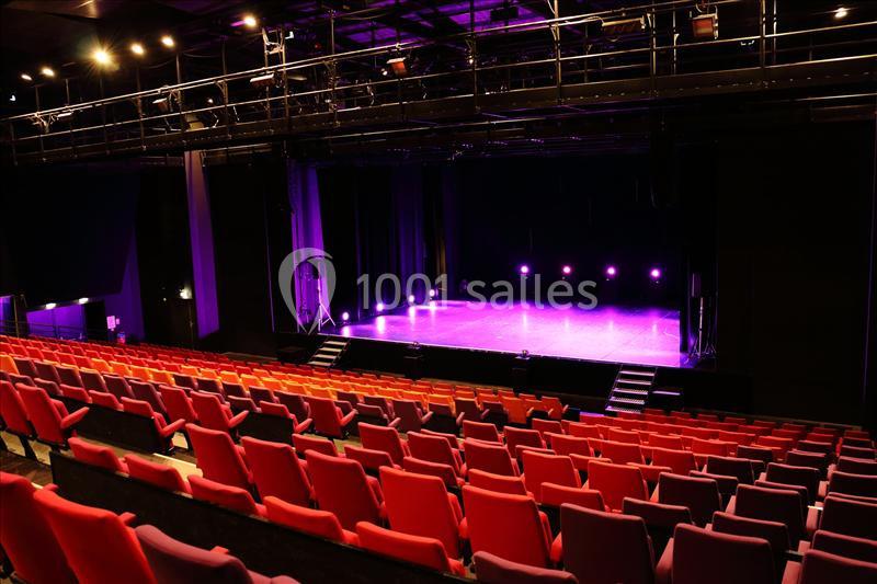 Salle de spectacle vide avec rangées de sièges rouges et scène éclairée par des projecteurs violets.