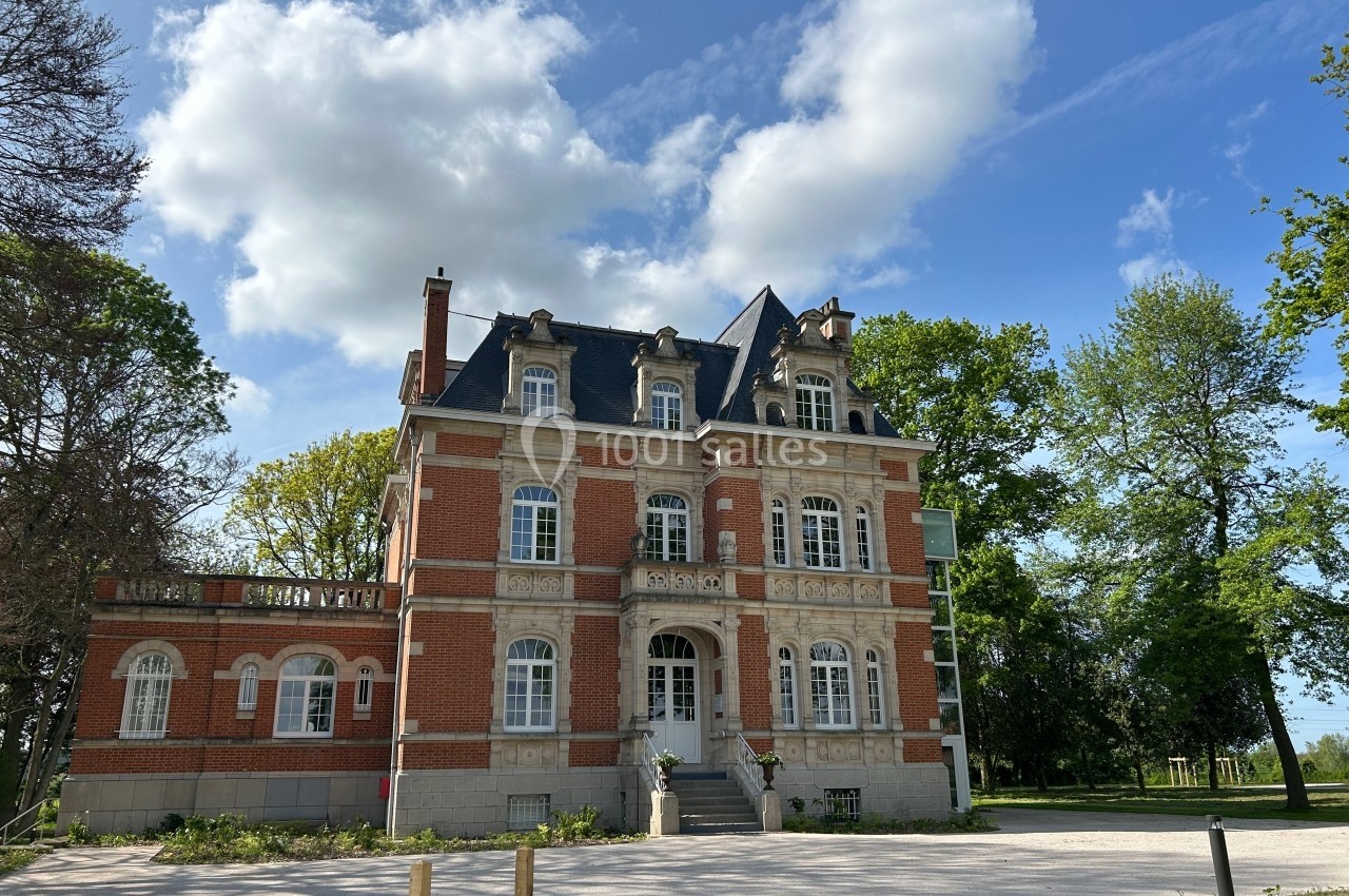 Façade d'un manoir en briques rouges avec toit en ardoise, entouré d'arbres sous un ciel partiellement nuageux.