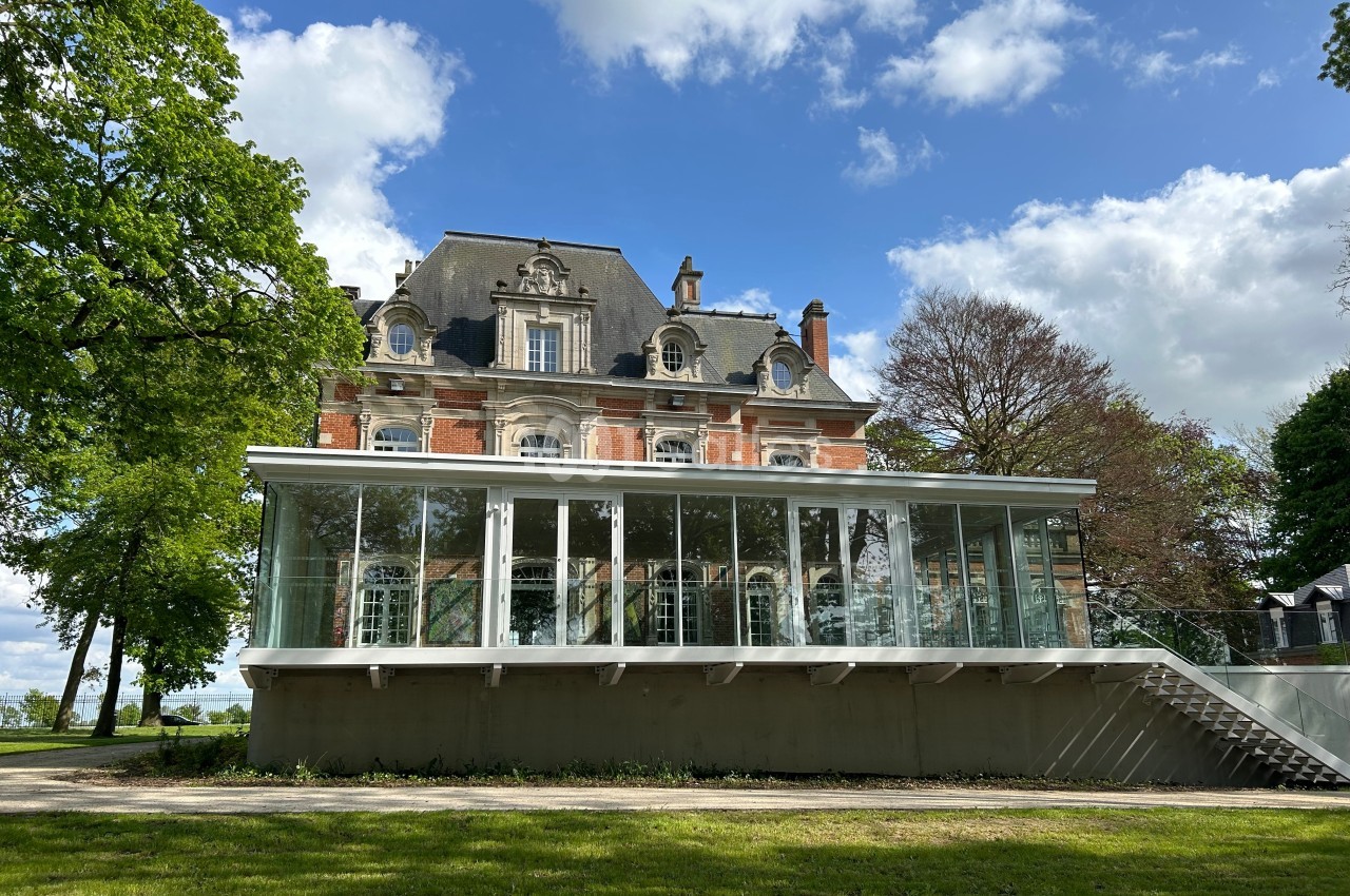 Façade d'un bâtiment ancien en briques rouges avec une extension moderne vitrée, entouré de verdure et d'arbres.