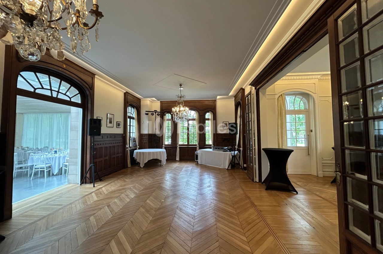 Salle élégante avec parquet en bois, lustres, grandes fenêtres et tables dressées visibles dans une pièce adjacente.