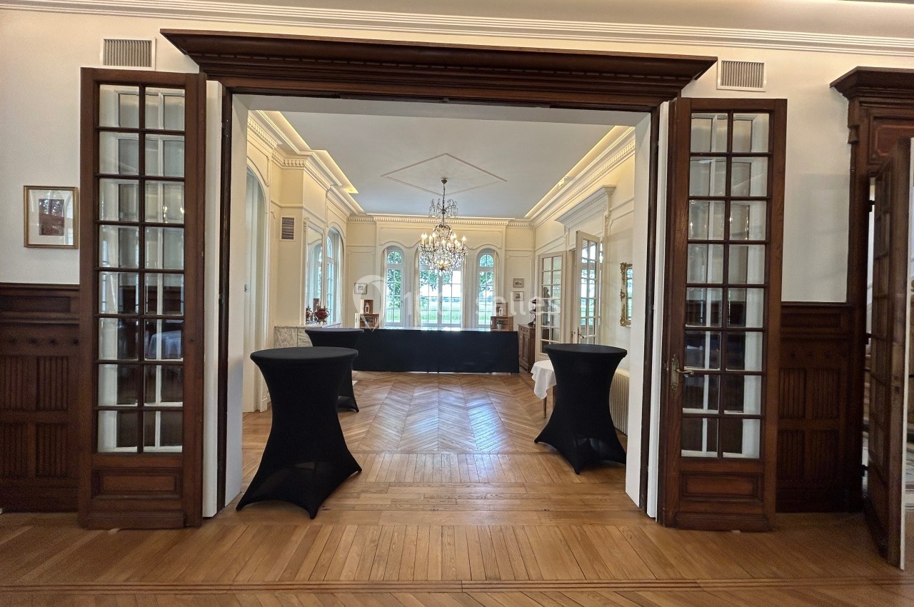Salle élégante avec parquet en bois, tables hautes noires et lustre suspendu, encadrée par des portes vitrées.