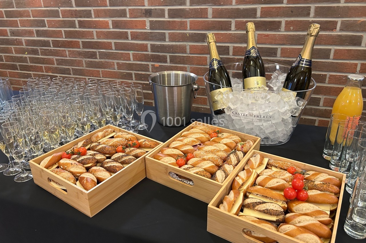 Plateau de pains variés avec tomates cerises, flûtes à champagne, bouteilles sur glace et jus d'orange sur une table.