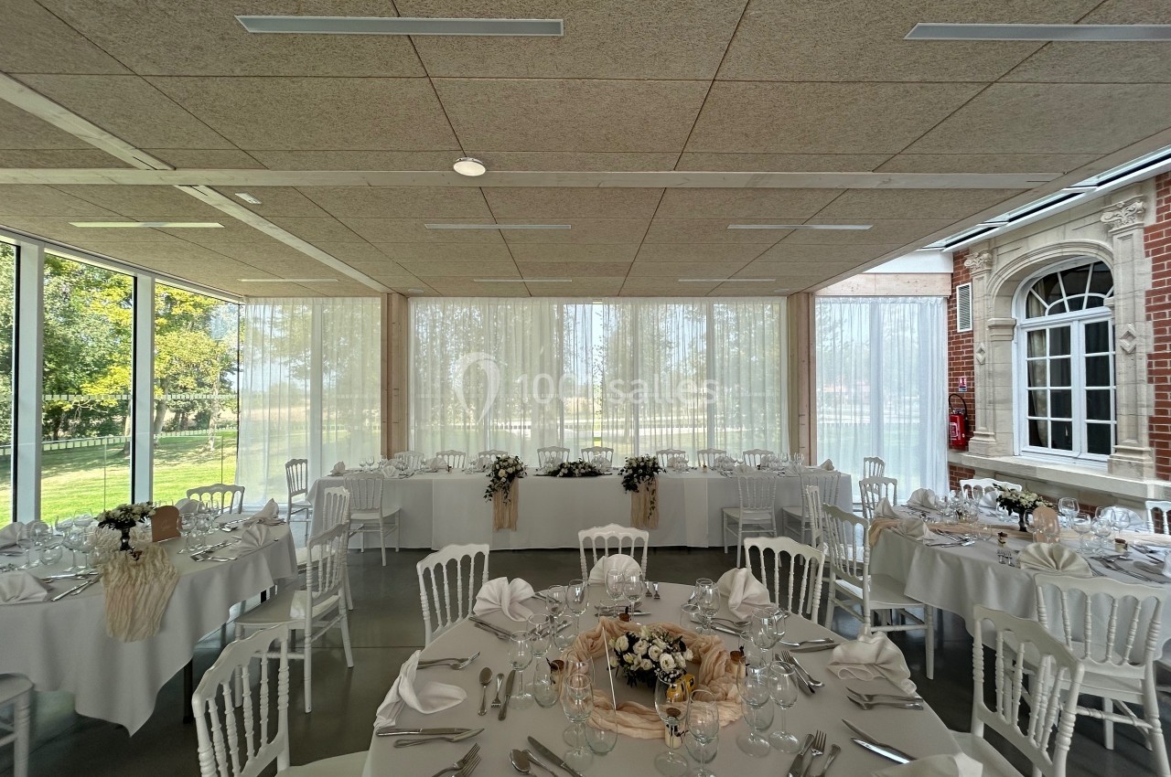 Salle de réception lumineuse avec tables rondes décorées pour un événement, vue sur un jardin à travers de grandes baies…