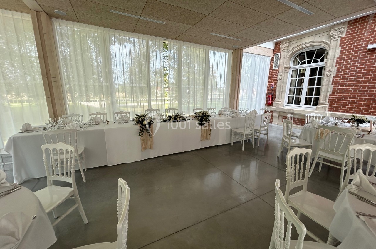 Salle de réception lumineuse avec tables décorées de nappes blanches, chaises assorties et grandes baies vitrées.