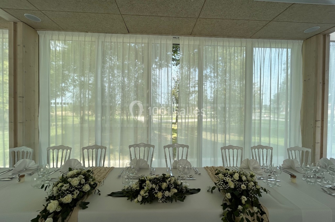Table décorée pour un événement, ornée de fleurs blanches et de verdure, devant de grandes fenêtres avec rideaux…