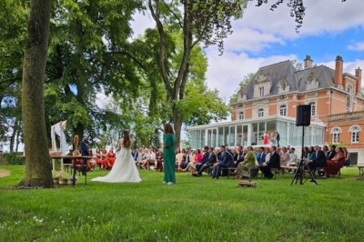 Cérémonie en plein air dans un parc, avec des invités assis face à une mariée et un officiant, devant un manoir.