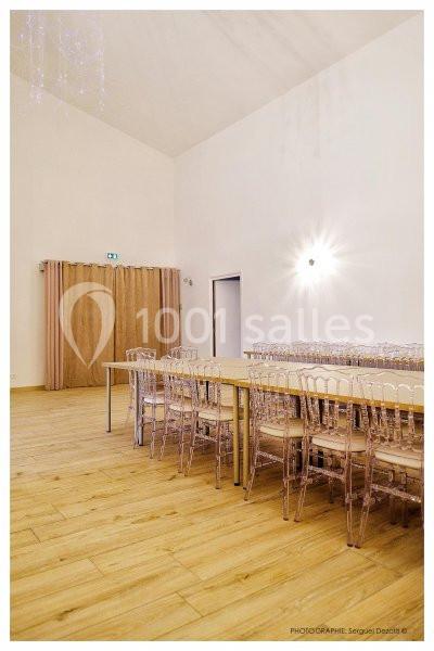 Salle lumineuse avec parquet, tables alignées et chaises transparentes, rideaux beige couvrant une porte au fond.