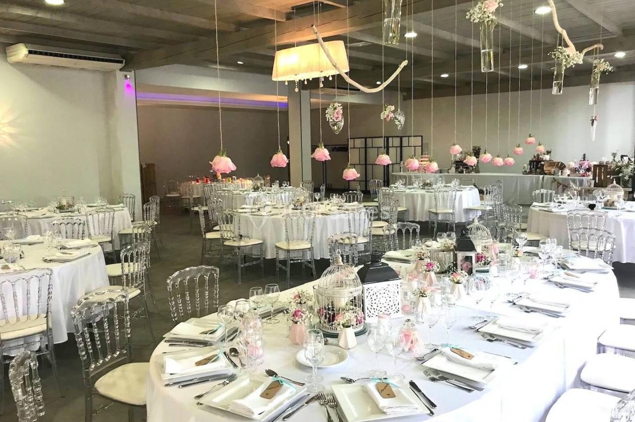 Salle de réception décorée avec des tables rondes dressées, des chaises transparentes et des suspensions florales.