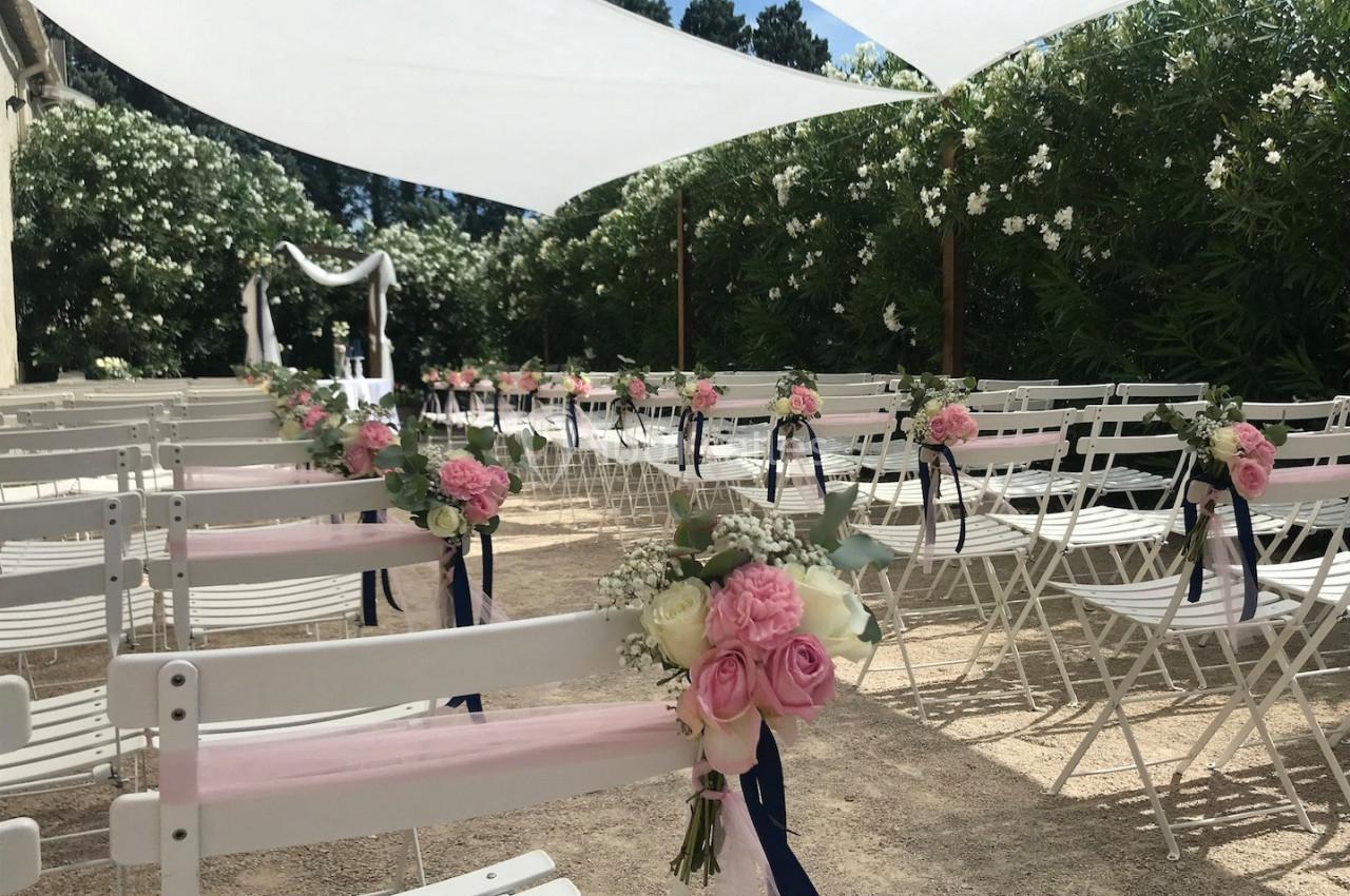 Chaises blanches décorées de fleurs roses et rubans, alignées pour une cérémonie en extérieur sous des voiles d'ombrage.