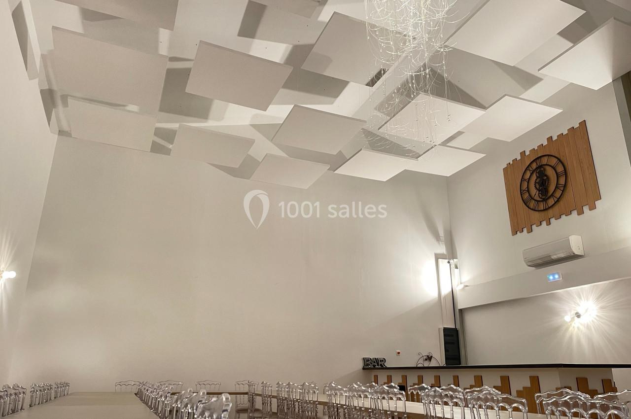 Salle de réception avec plafond acoustique, lustre moderne, horloge murale et chaises transparentes alignées.