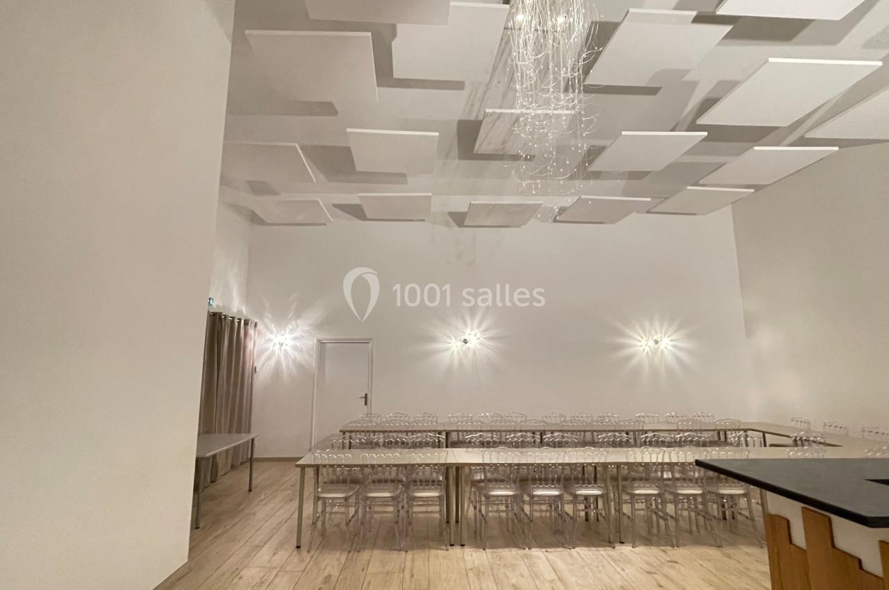 Salle lumineuse avec plafond décoratif, tables alignées avec chaises transparentes et éclairage mural discret.