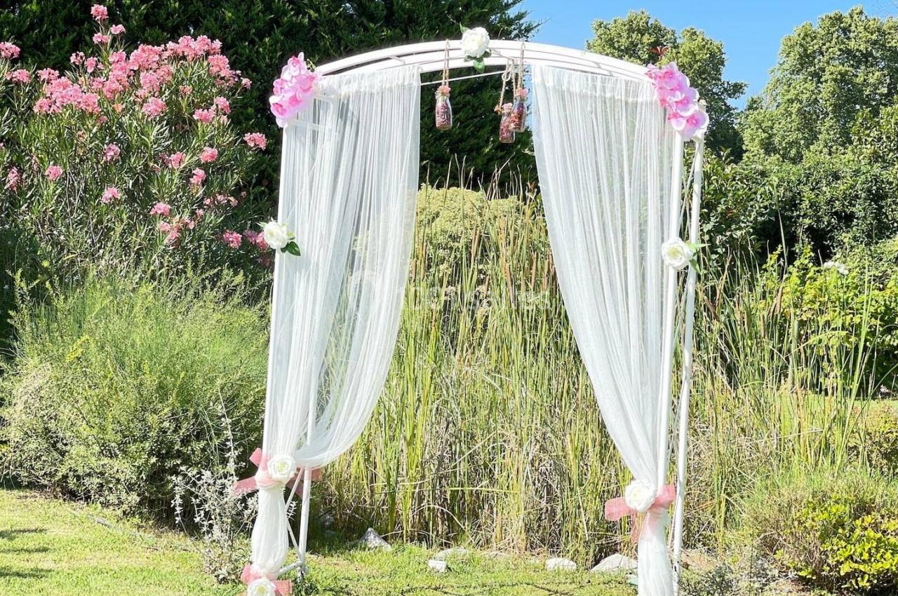 Arche de cérémonie décorée de voilages blancs et de fleurs roses, installée dans un jardin verdoyant.