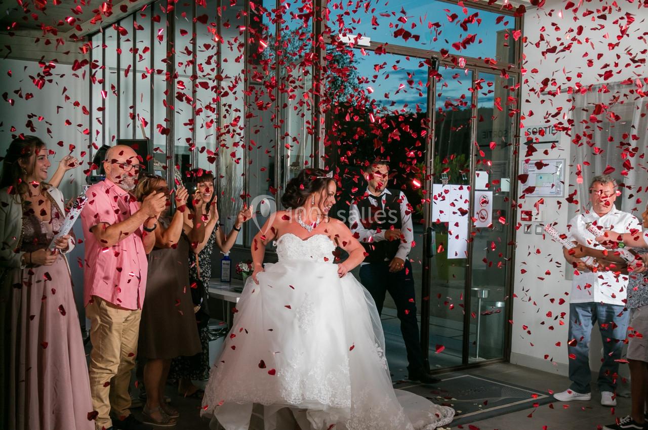 Une mariée en robe blanche sort d'un bâtiment sous une pluie de pétales rouges, entourée d'invités applaudissant.