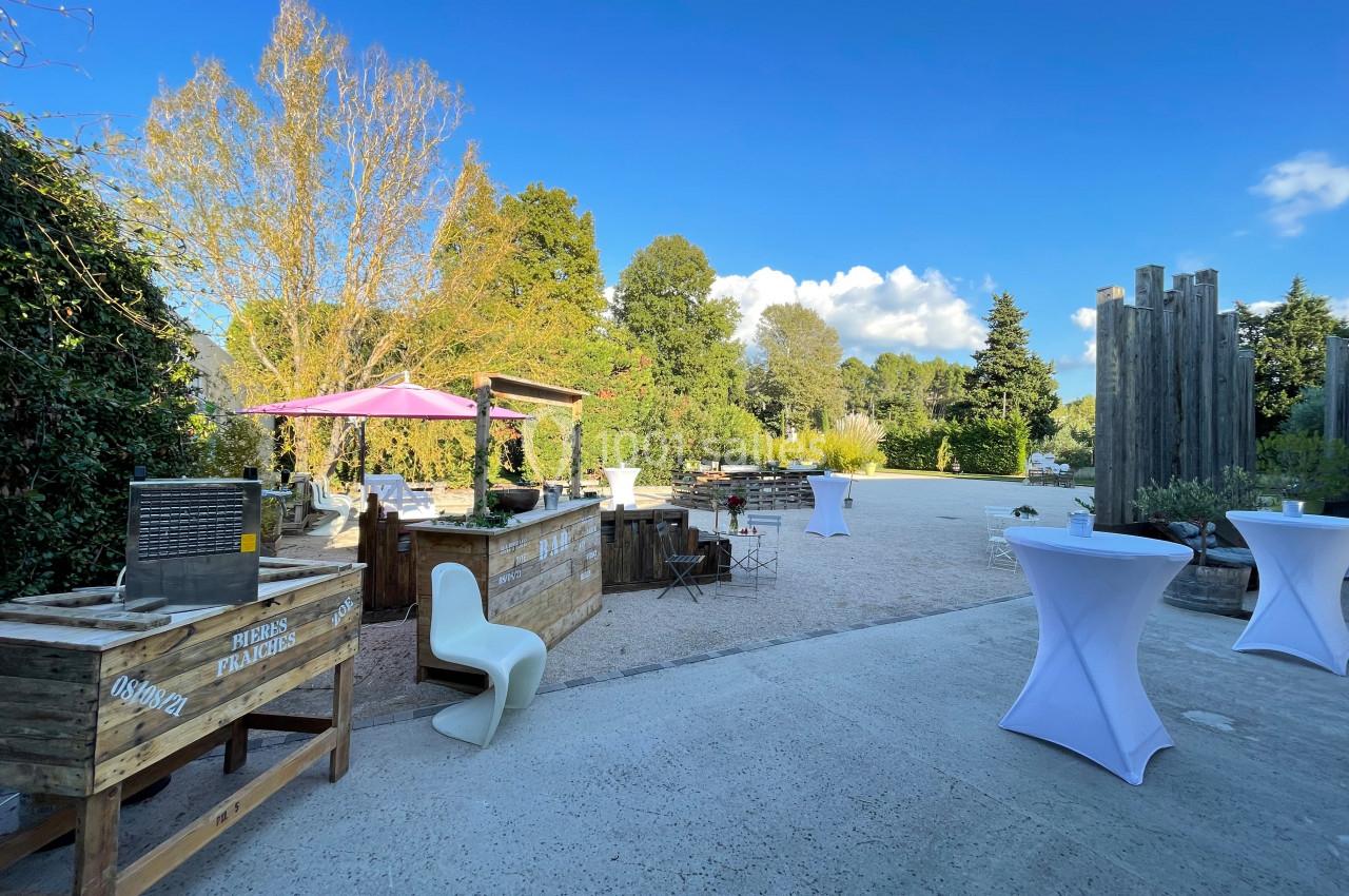 Espace extérieur avec tables hautes, chaises design et stands en bois, entouré de végétation sous un ciel dégagé.
