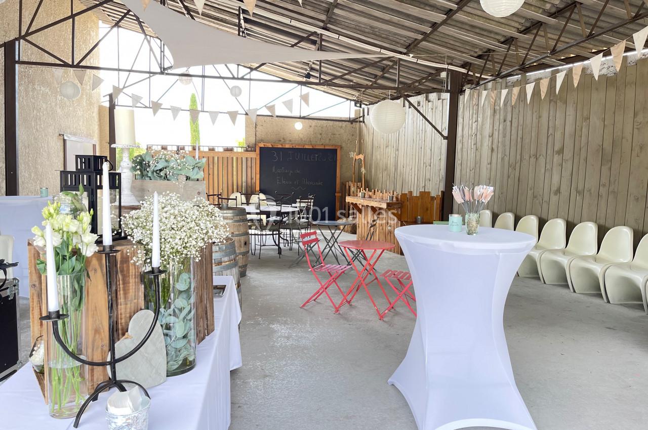 Espace extérieur décoré pour un événement, avec guirlandes, tables hautes, chaises et fleurs.