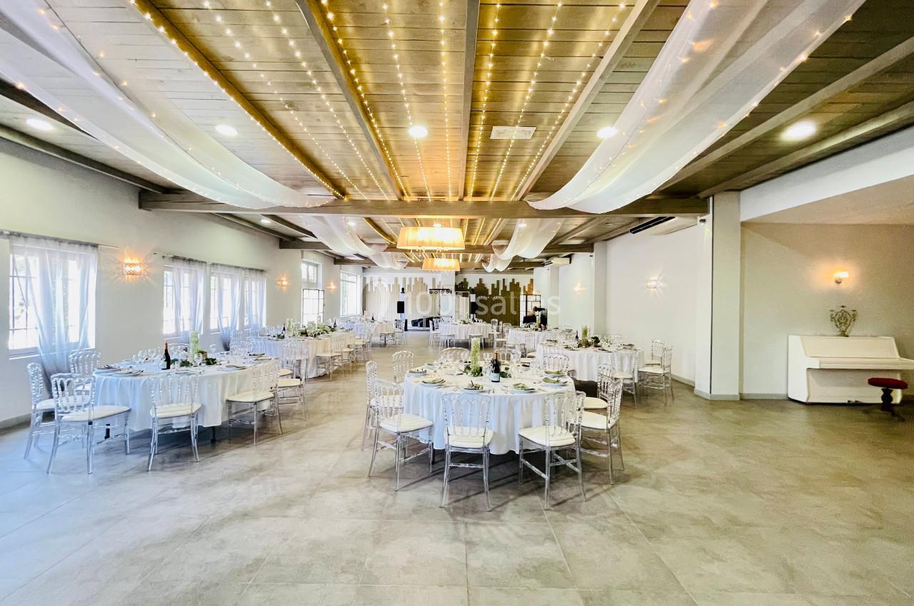 Salle de réception lumineuse avec tables rondes dressées, décorations suspendues et éclairage chaleureux.