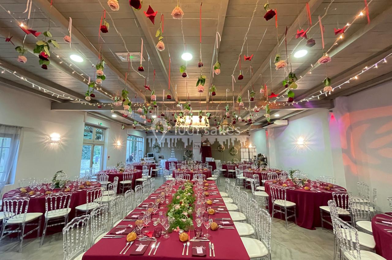 Salle de réception décorée avec des tables dressées, nappes bordeaux et fleurs suspendues au plafond.