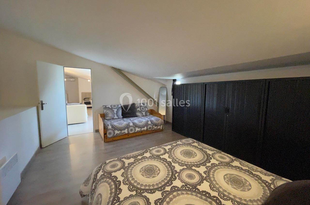 Chambre mansardée avec lit double, canapé, armoire noire et porte ouverte donnant sur une autre pièce.