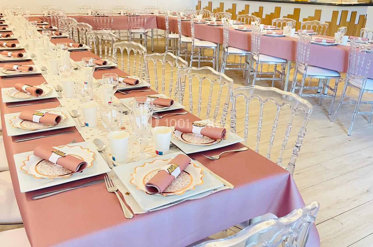Tables dressées avec nappes roses, vaisselle élégante et chaises transparentes dans une salle de réception lumineuse.