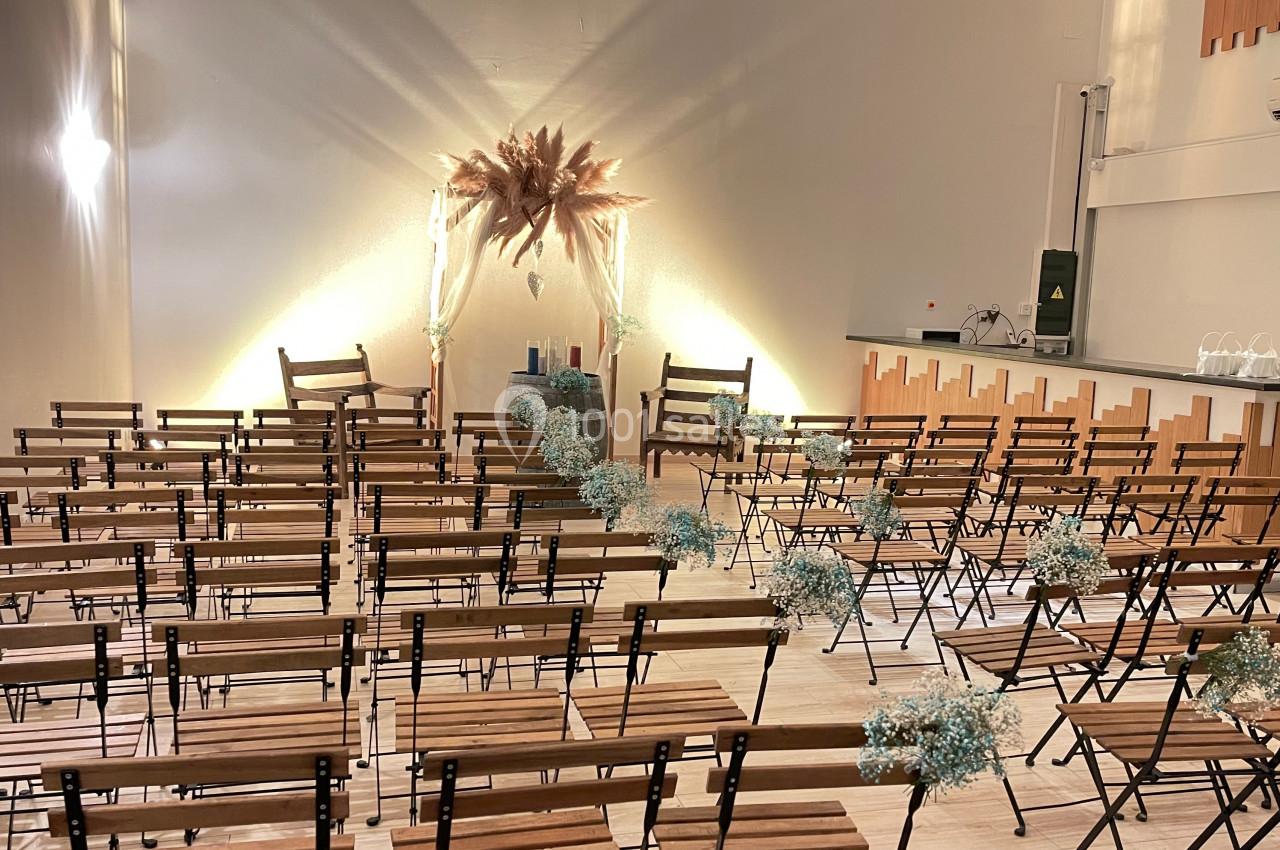 Salle décorée pour une cérémonie avec des chaises en bois alignées, des fleurs séchées et un arrière-plan lumineux.