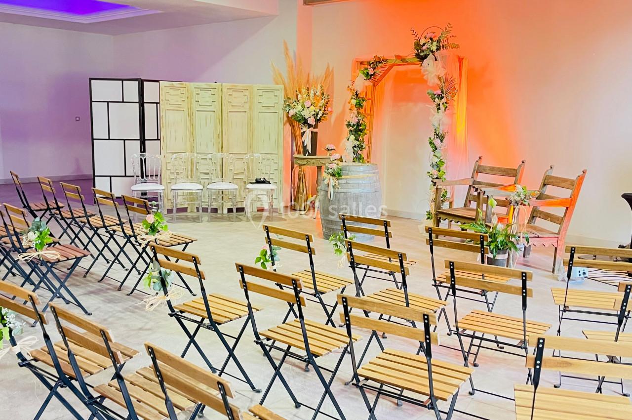 Salle décorée pour une cérémonie, avec des chaises en bois alignées, une arche fleurie et un éclairage chaleureux.
