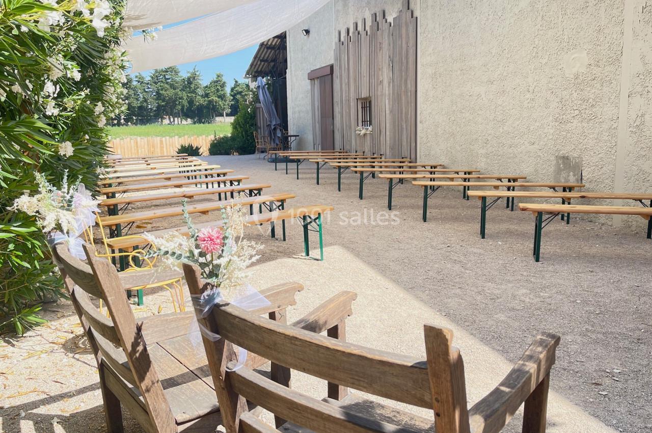 Chaises et bancs en bois disposés en extérieur pour une cérémonie, sous des voiles d'ombrage près d'un mur clair.