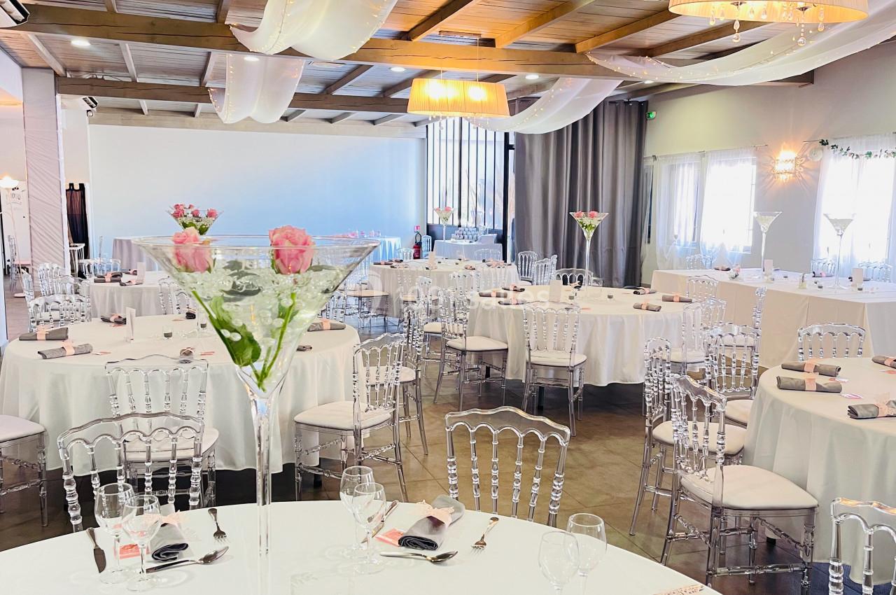 Salle de réception décorée avec des tables rondes, nappes blanches, chaises transparentes et centres de table fleuris.