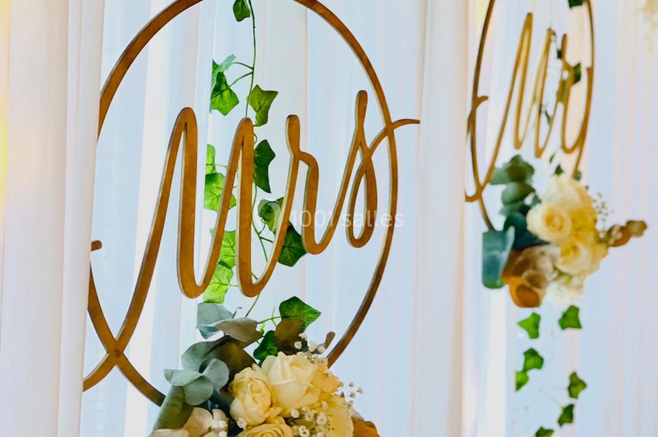 Anneaux décoratifs dorés avec l'inscription ’Mrs’, ornés de fleurs et de feuillage, suspendus devant un rideau blanc.