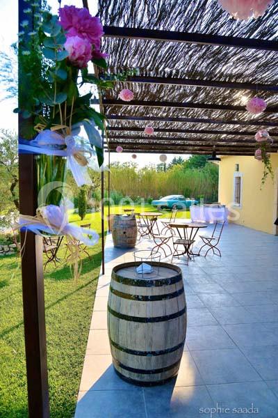 Terrasse ombragée avec tables en métal, tonneau en bois en premier plan, et jardin verdoyant en arrière-plan.