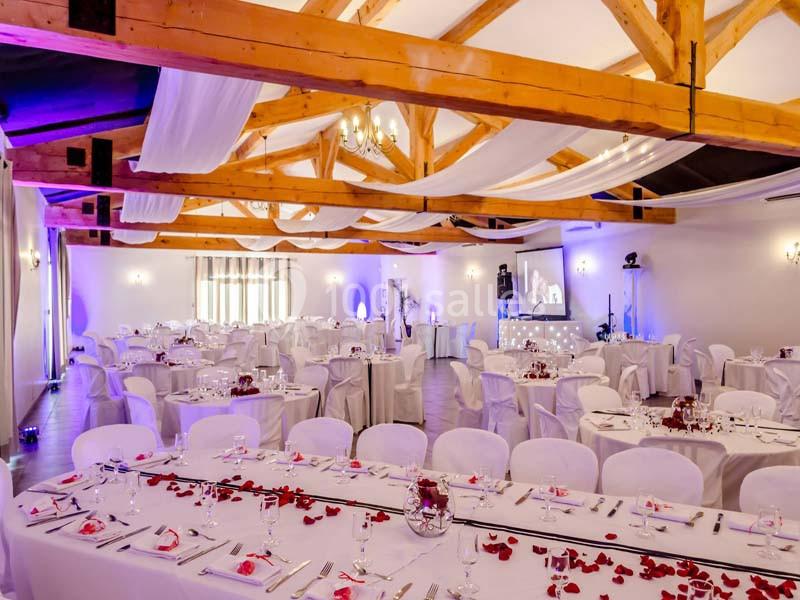 Salle de réception décorée avec des tables dressées, nappes blanches, pétales rouges et poutres apparentes en bois.