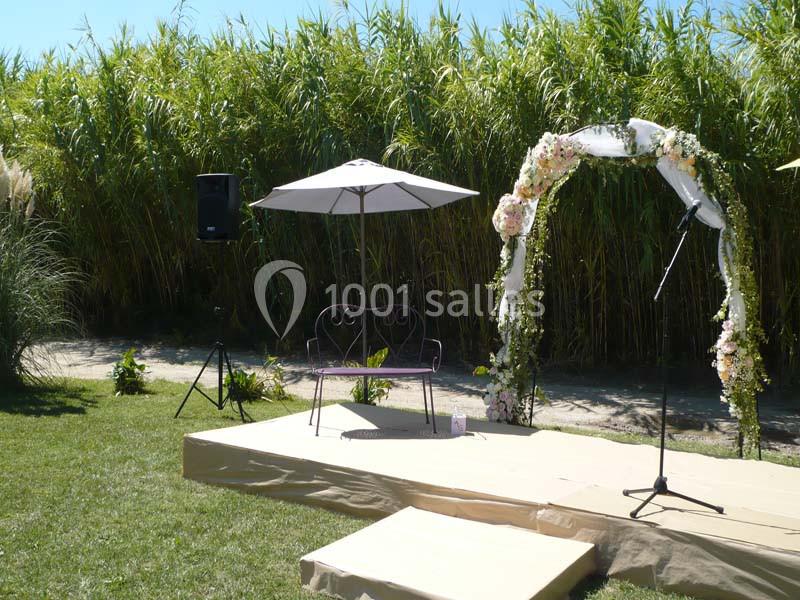 Estrade en extérieur avec arche fleurie, parasol, banc en métal et micro, entourée de végétation dense.
