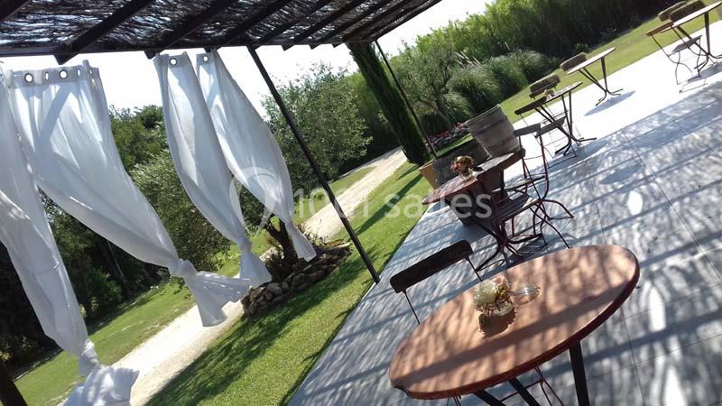 Terrasse extérieure avec tables, chaises et rideaux blancs flottants, entourée de verdure et d'arbres.