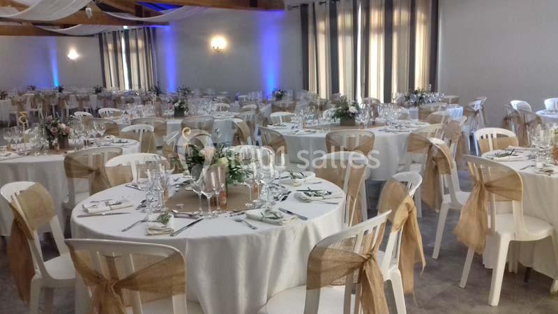 Salle de réception décorée avec des tables rondes dressées, nappes blanches et chaises ornées de rubans beige.
