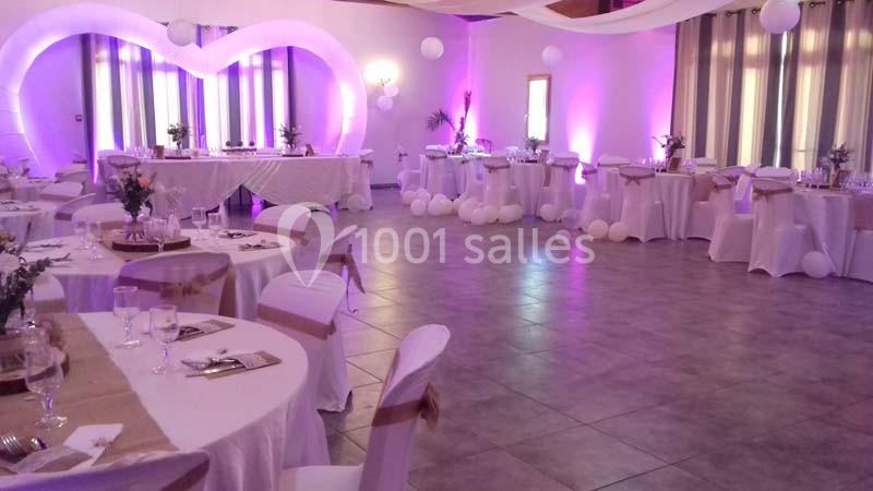 Salle de réception décorée avec des tables rondes, nappes blanches, ballons et éclairage violet.