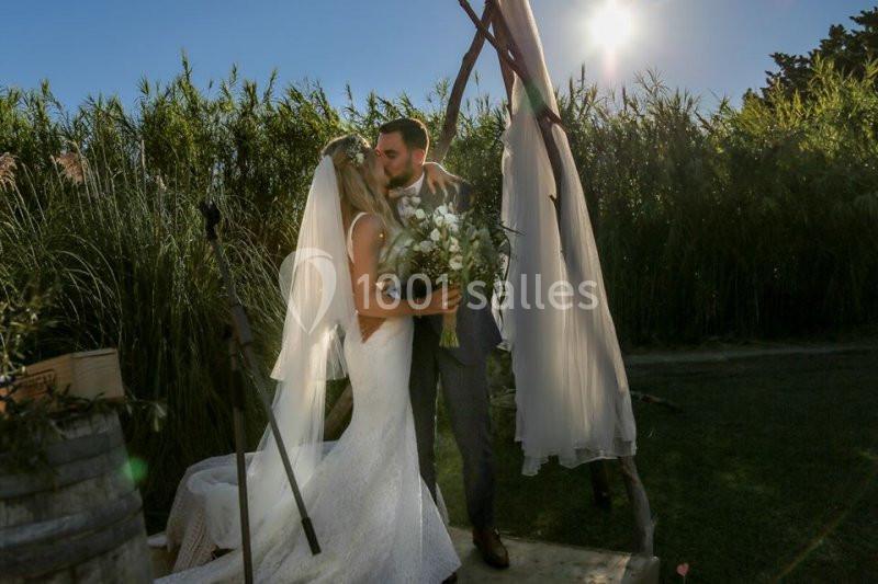 Un couple en tenue de mariage s'embrasse sous une arche décorée, entouré de verdure et éclairé par le soleil.