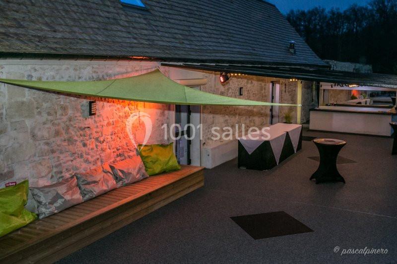 Terrasse extérieure en soirée avec banc en bois, coussins, voile d'ombrage et tables hautes près d'un mur en pierre.