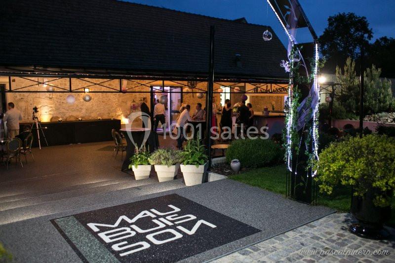 Entrée d'un lieu événementiel en soirée, avec terrasse éclairée, plantes en pot et invités en arrière-plan.