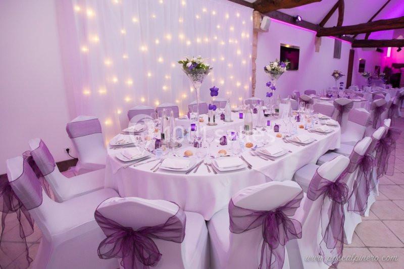 Salle de réception décorée avec des tables rondes, nappes blanches, chaises ornées de nœuds violets et guirlandes lumineuses.