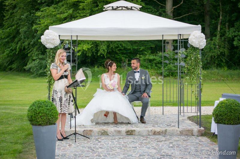 Un couple assis sous une tonnelle blanche lors d'une cérémonie de mariage en plein air, avec une officiant à gauche.