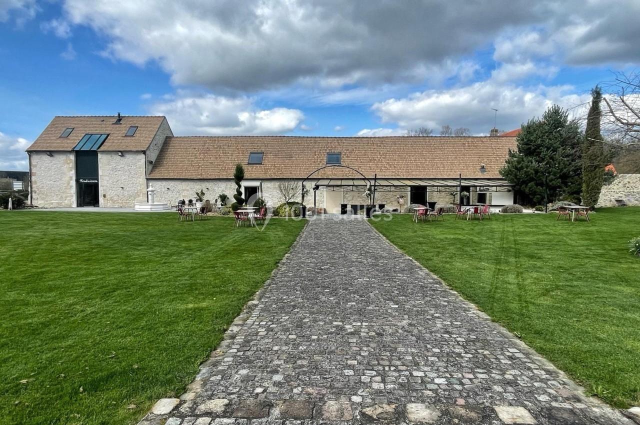 Allée pavée menant à un bâtiment en pierre avec toit en tuiles, entouré de pelouse et de quelques tables extérieures.