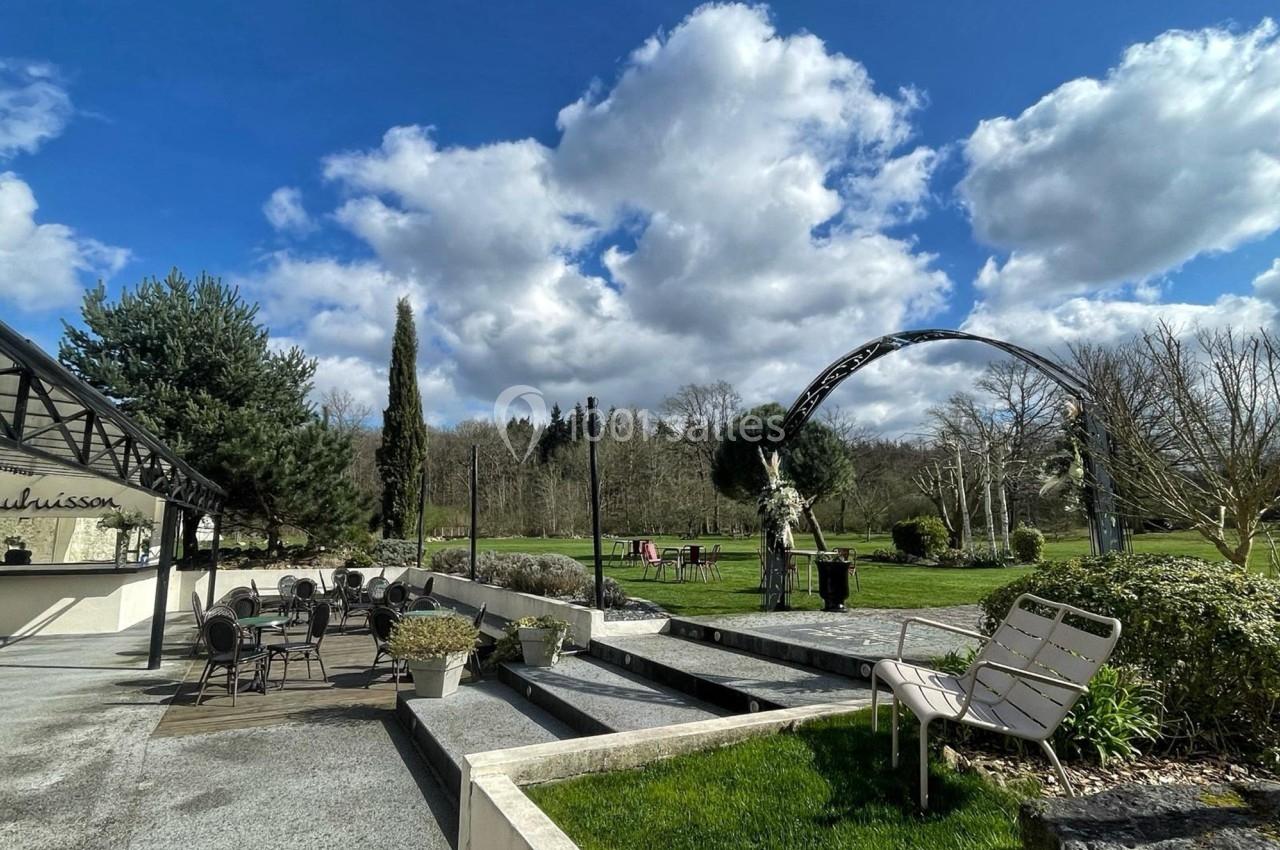 Terrasse extérieure avec tables et chaises, jardin verdoyant, arche décorative et ciel partiellement nuageux.